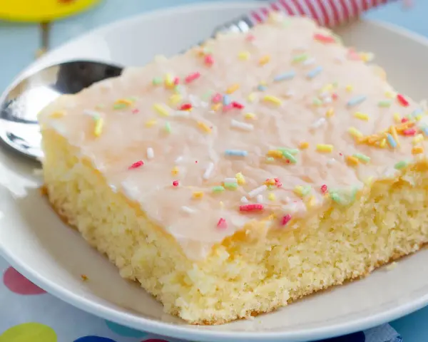 Ein Stück saftiger Fantakuchen vom Blech, mit Zuckerguss und bunten Streuseln bestreut, auf einem Teller mit Löffel.