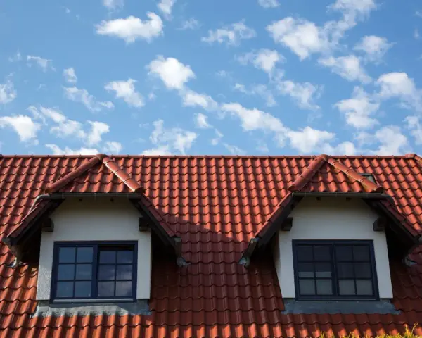 Czerwony dach z dachówki ceramicznej, z dwoma lukarnami i niebieskim niebem w chmurach.