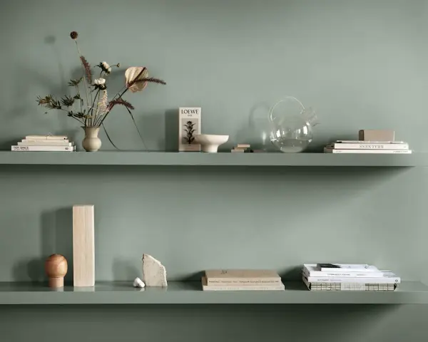 Två gröna hyllplan med böcker, vas med blommor, glasburk och dekorativa föremål. Enkel och stilren inredning.
