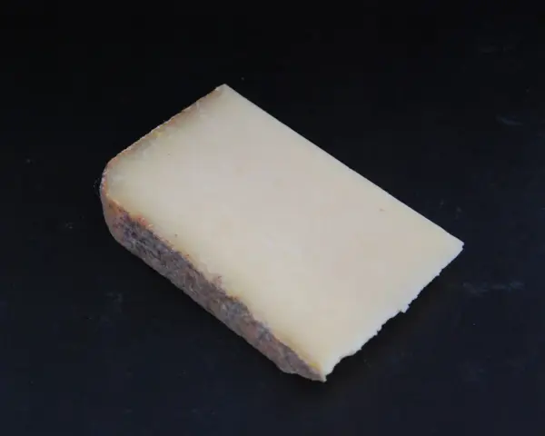 Un morceau de Cantal, lait cru ou pasteurisé, à la croûte rustique et à la pâte jaune pâle, posé sur un fond sombre.