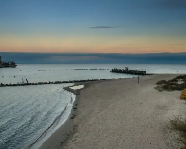 Zachód słońca nad morzem w Gdyni. Na wodzie widać betonową fortecę, a na brzegu piaszczystą plażę i babie doły.