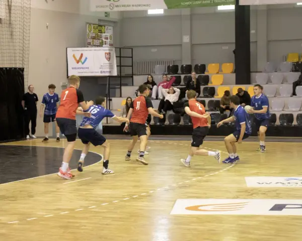 Kacper Wachowiak w akcji podczas meczu piłki ręcznej. Zawodnicy w czerwonych i niebieskich strojach walczą o piłkę na parkiecie.