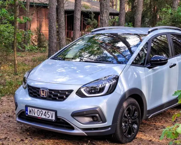 Jasny Honda Jazz Crosstar zaparkowany w lesie, obok drewnianego domku.