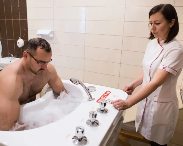 Mężczyzna w wannie z pianą, terapeuta przygotowuje masaż wirowy kończyn dolnych.