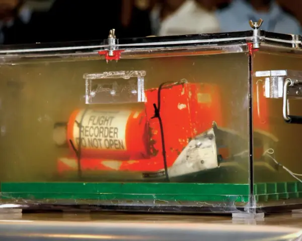 Pomarańczowa skrzynka "czarnej skrzynki" z napisem "FLIGHT RECORDER DO NOT OPEN" w przezroczystej obudowie, dowód w sprawie wypadku lotniczego.