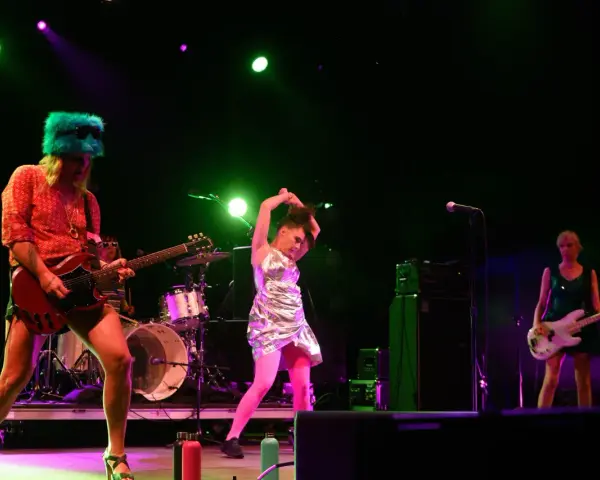 Sur scène, le groupe Bikini Kill enflamme le public. Une guitariste porte un chapeau vert fluo, une autre joue de la basse, et une troisième danse avec énergie.