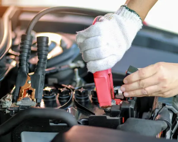 Eine Hand mit Handschuh klemmt das rote Kabel an den Pluspol, während die andere Hand den Minuspol der Autobatterie sichert.
