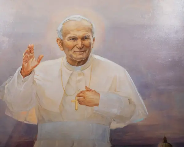 Obraz przedstawia Jana Pawła II w białej szacie, z podniesioną prawą dłonią i krzyżem na piersi. Wierni odmawiają modlitwę do Jana Pawła II o uzdrowienie.