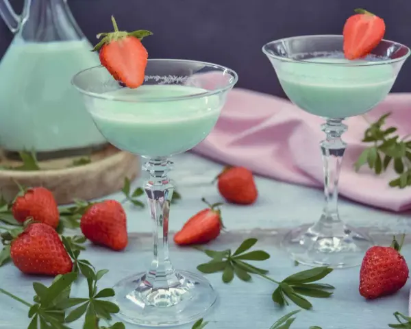 Zwei Gläser mit hellgrünem Likör, garniert mit Erdbeeren. Ein Krug mit dem Likör und frische Erdbeeren deuten darauf hin, dass man Waldmeister Schnaps selber machen kann.