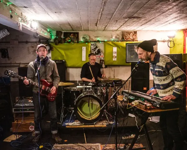 Un groupe de musique punk des années 90 joue dans un local sombre. Un guitariste, un batteur et un claviériste sont sur scène.