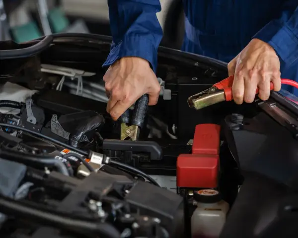Hände mit Starthilfekabeln, um die **Batterie richtig anzuklemmen**. Ein Mechaniker in blauer Arbeitskleidung verbindet die Klemmen mit der Autobatterie.