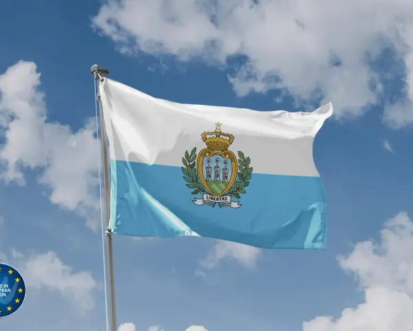 Flaga San Marino powiewa na tle błękitnego nieba z chmurami. Herb z trzema wieżami i koroną zdobi biało-błękitny materiał.