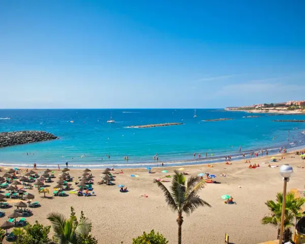 Spiaggia affollata con ombrelloni e lettini, mare azzurro e città costiera sullo sfondo. Sembra l'isola più bella delle Canarie.
