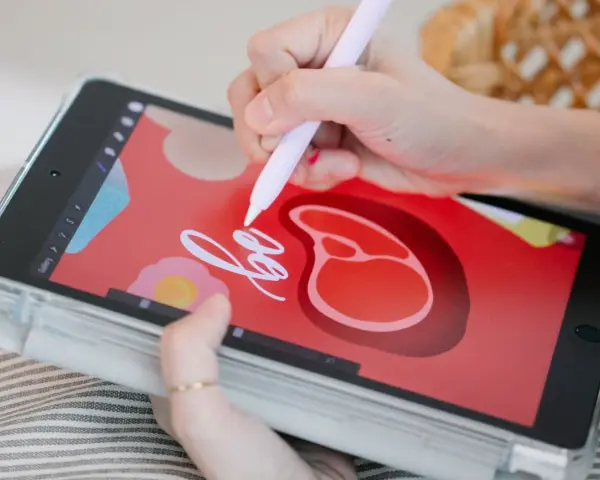 Rysowanie na tablecie graficznym bez podłączania do komputera. Dłoń z rysikiem tworzy napis "be" na czerwonym tle z motywami jedzenia.