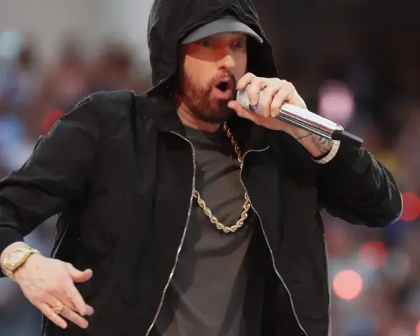 Eminem w czarnej bluzie z kapturem śpiewa do mikrofonu, w złotym łańcuchu i zegarku.