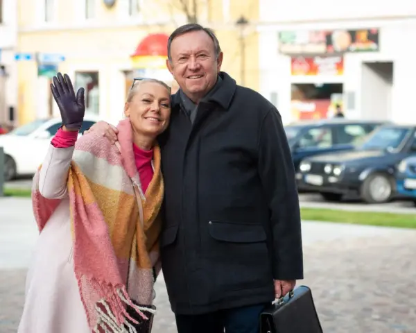 Małgorzata Szeptycka wita się z uśmiechem, obejmując partnera. W tle miejski krajobraz z samochodami.