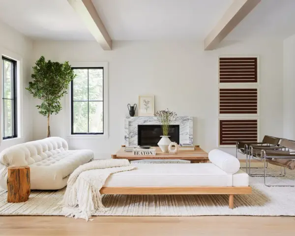 Un elegante salón blanco y madera natural, con sofá curvo, chaise longue, chimenea de mármol y sillas de cuero.