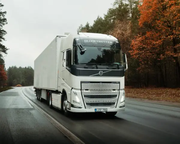 Biała ciężarówka Volvo FH12 z naczepą jedzie po mokrej drodze wśród jesiennego lasu.