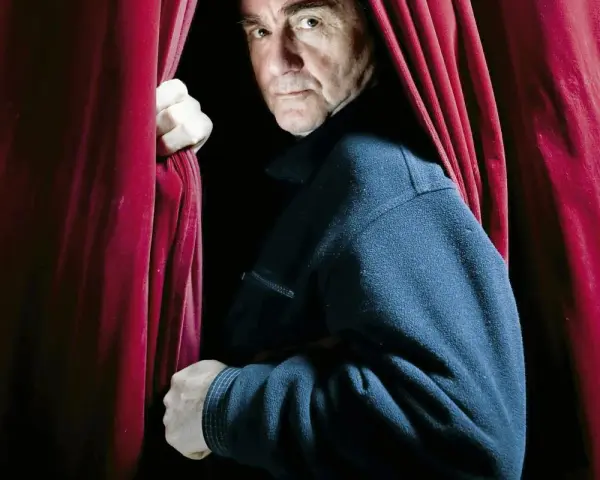 Jan Peszek młody, wychodząc zza czerwonej kurtyny, spogląda wprost na widza.