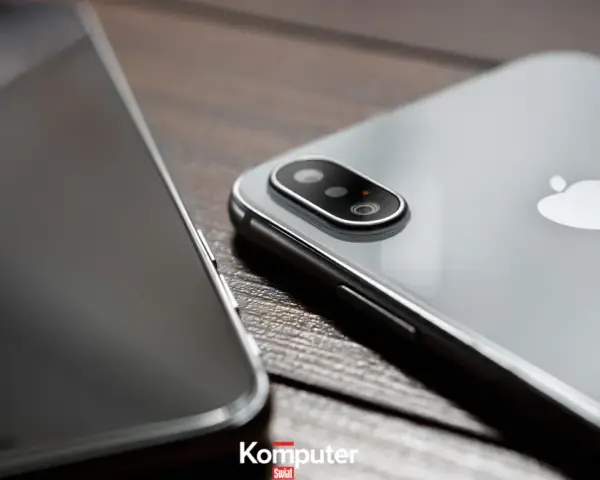 Dwa telefony leżą obok siebie na drewnianym blacie. Jeden z nich, srebrny iPhone, ma widoczny moduł aparatu. Drugi, z ciemnym ekranem, jest ustawiony bokiem. Idealne do rozmowy przez **Facetime**.