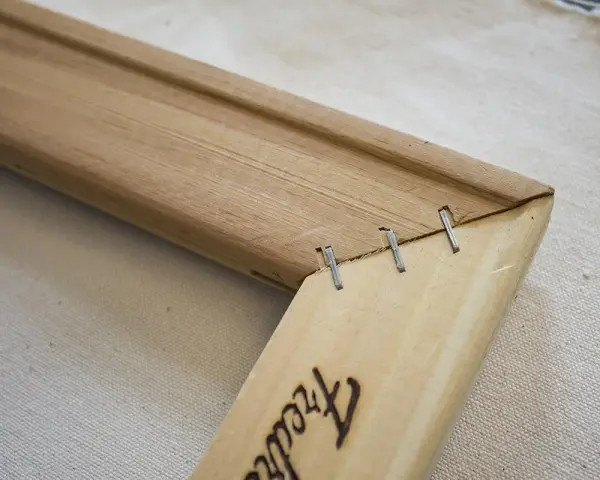 Détail d'un coin de cadre en bois, montrant comment tendre une toile sans châssis avec des agrafes.