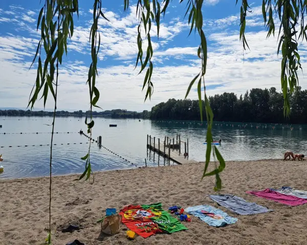 Ein sonniger Tag am Lauterbourg See. Handtücher liegen am Sandstrand, Kinder planschen im Wasser.