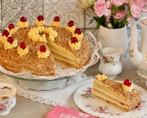 Ein köstlicher Frankfurter Kranz, verziert mit Buttercreme-Tupfern und Kirschen, umgeben von gehackten Nüssen. Ein Stück ist auf einem Teller angerichtet.