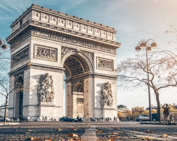La majestuosa entrada del Arco del Triunfo en París, con esculturas y detalles ornamentales, bajo un cielo azul y árboles otoñales.