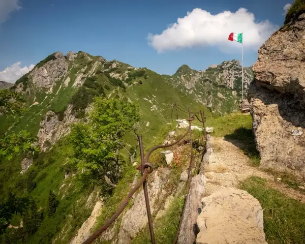 Sentiero della Pace tra montagne verdi, con bandiera italiana che sventola. Un percorso che ricorda la storia.