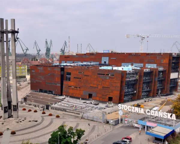 Nowoczesne muzeum na terenie Stoczni Gdańskiej, z charakterystycznymi, rdzawymi fasadami, w otoczeniu dźwigów stoczniowych i Pomnika Poległych Stoczniowców.