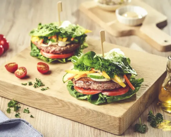 Dwa soczyste burgery w sałacie, z pomidorami, ogórkiem i papryką, na drewnianej desce.