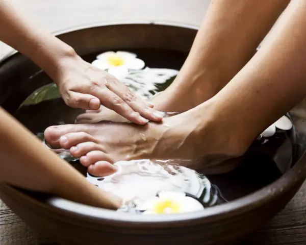 Soaking feet in a warm bath with Epsom salt for feet infection. A hand gently massages the foot, surrounded by water and white flowers.