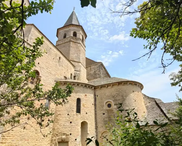 Vue d'une église romane à Villeneuve de Rouergue, encadrée par des arbres verdoyants sous un ciel bleu parsemé de nuages.