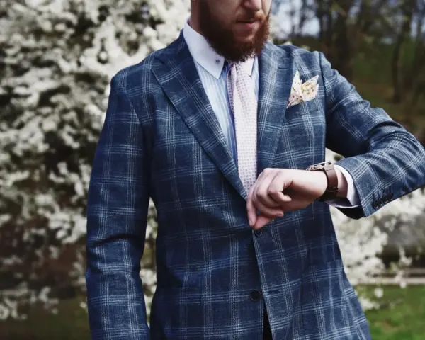 Elegancki mężczyzna w garniturze w kratę, z akcentem w kolorze turkusowym w butonierce, sprawdza zegarek.
