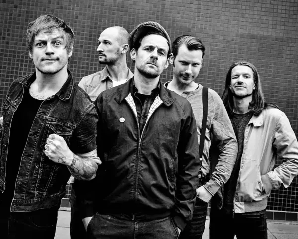 Fünf Männer stehen vor einer gemusterten Wand. Sie sehen aus wie eine energiegeladene Punkband, bereit, die Bühne zu rocken.