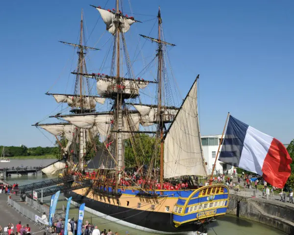 Le majestueux bateau l'Hermione, réplique d'une frégate du XVIIIe siècle, est amarré, pavoisé du drapeau français. Une foule s'est rassemblée pour admirer ce navire historique.