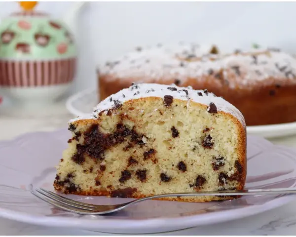 Una fetta di soffice torta con gocce di cioccolato, cosparsa di zucchero a velo, su un piatto lilla con una forchetta.