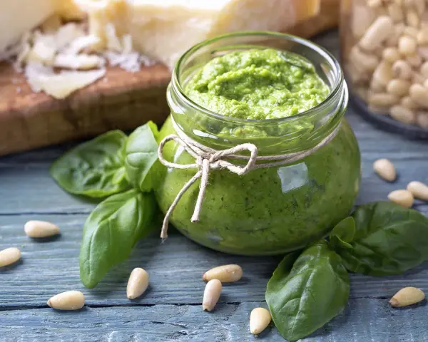 Słoik domowego pesto z bazylii, otoczony świeżymi liśćmi bazylii i orzeszkami pinii. Obok kawałki parmezanu. Idealne do makaronu.