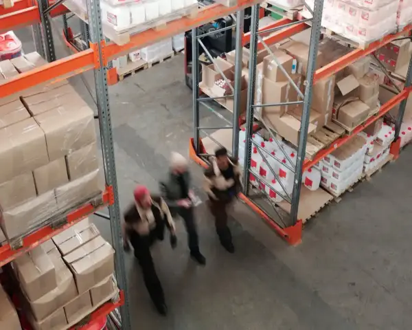 Magazzino con scaffalature piene di scatoloni e taniche. Tre persone camminano velocemente, forse per svolgere mansioni di aiuto magazziniere.