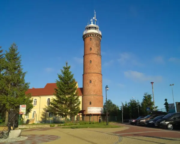 Wysoka, ceglana latarnia morska Jarosławiec góruje nad placem z drzewami i budynkiem.