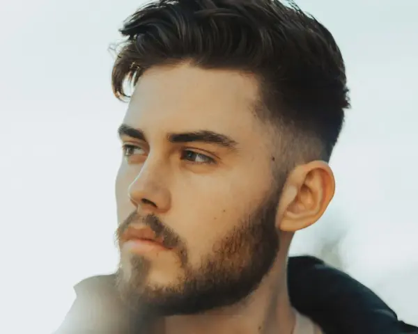 Ein Mann mit Bart und einem modernen taper fade design im Haar blickt nachdenklich zur Seite.