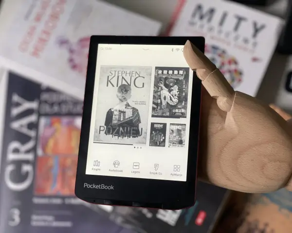 Drewniana dłoń wskazuje na ekran czytnika PocketBook, gdzie wyświetla się okładka książki Stephena Kinga "Później". W tle widać inne książki.