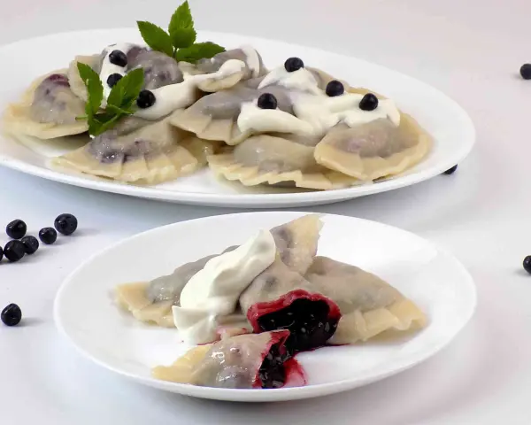 Pyszne pierogi z jagodami, polane śmietaną i ozdobione miętą. Na talerzu widać też kilka świeżych jagód.