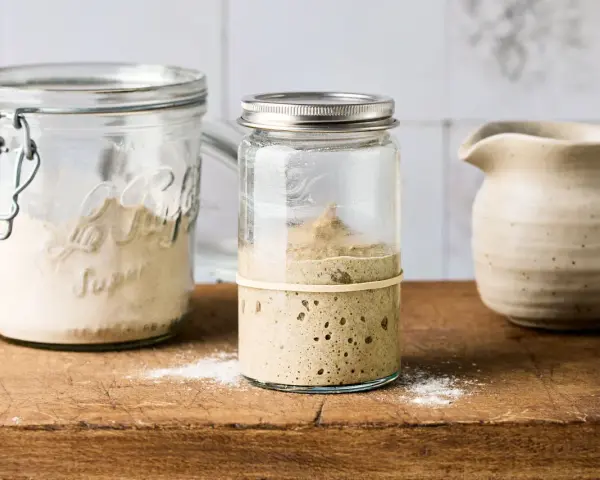 Blubbernder Sauerteig im Glas, bereit für schnelles Sauerteig herstellen. Daneben ein Glas mit Mehl und ein Krug.