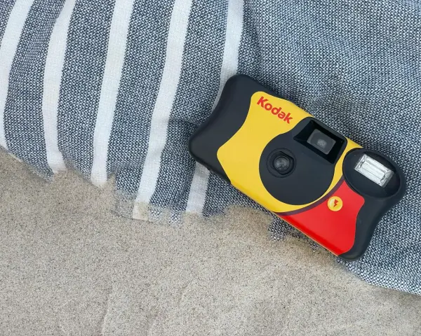 Aparat jednorazowy firmy Kodak leży na plaży obok ręcznika. Idealny do uchwycenia letnich wspomnień.