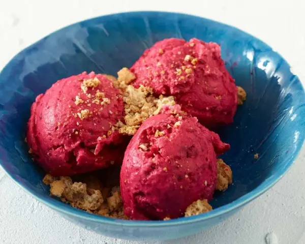 Drei Kugeln leuchtend pinkes Sauerrahm Eis, bestreut mit knusprigen Krümeln, in einer blauen Schale.