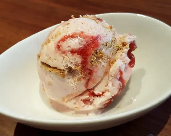 Ein Löffel cremiges strawberry cheesecake eis mit Erdbeersauce und Keksstückchen in einer weißen Schale.