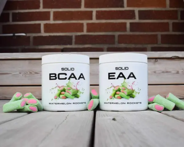 Två burkar med kosttillskott, en märkt BCAA och en EAA, står framför en tegelvägg. Runt burkarna ligger gröna godisar med rosa inuti, som ser ut som vattenmelonraketer.