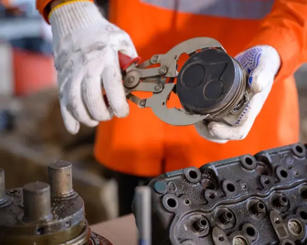 Mechanik w białych rękawicach trzyma tłok z widocznymi objawami zużytych pierścieni tłokowych. Obok leży głowica silnika.