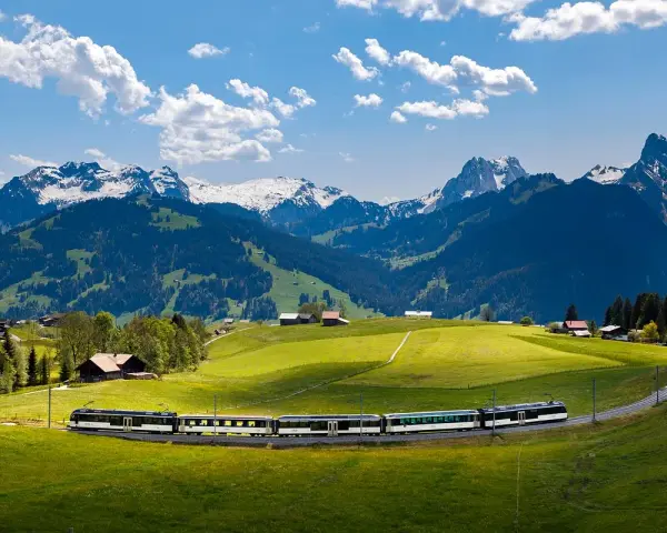 A train travels through a lush green valley with snow-capped mountains in the background. Many positive goldenpass line reviews mention this scenic route.